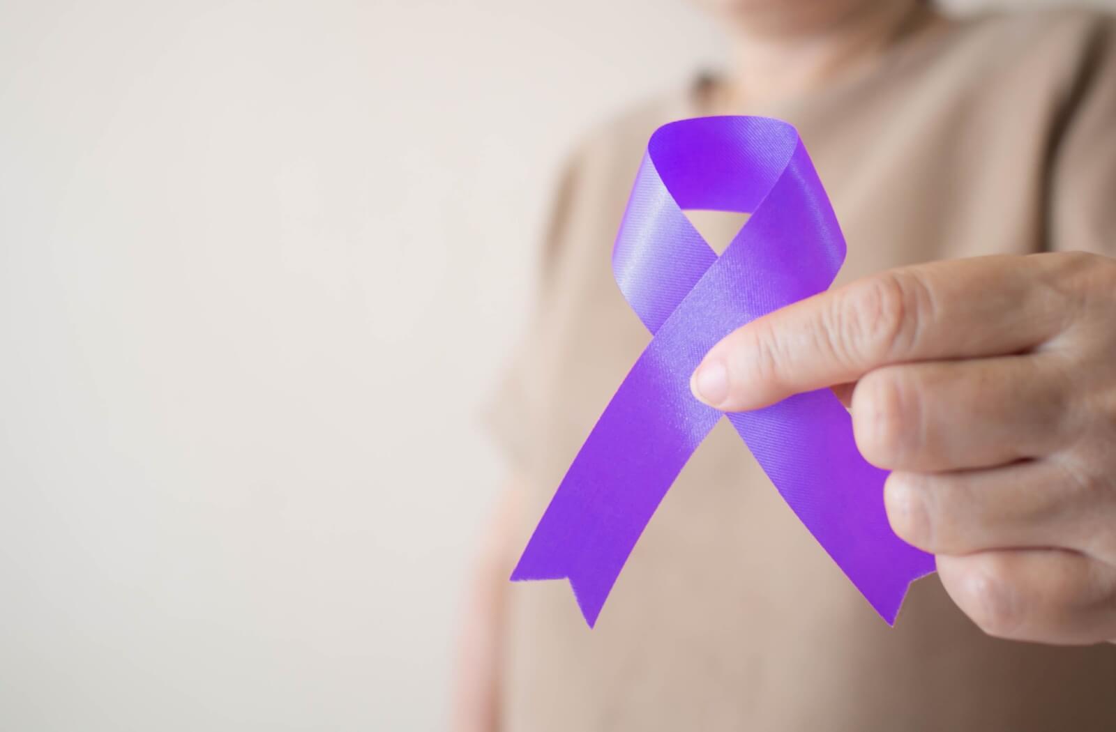 Against a grey wall, a senior holds out a glossy purple ribbon, which represents awareness for dementia